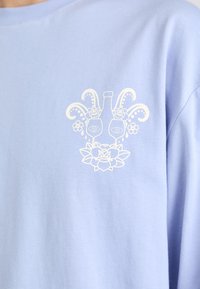 Light blue cotton t-shirt featuring a white graphic print of a bottle, eyes, and floral designs centered on the chest.