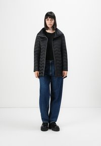 Vrouw in een zwarte gewatteerde jas over een zwart shirt, losse blauwe jeans en zwarte plateauschoenen, staande tegen een effen witte achtergrond.