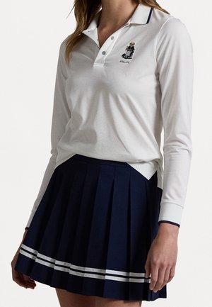 Mujer que lleva un polo blanco de manga larga con logotipo de oso y una falda plisada azul marino con dos franjas blancas cerca del dobladillo.