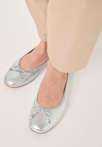 Ballerine argento con punta tonda, un piccolo fiocco sulla parte anteriore e una superficie liscia e lucida. I pantaloni beige sono parzialmente visibili.