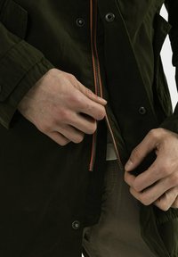 Manos subiendo la cremallera de una chaqueta verde oscuro con botones de presión negros visibles sobre pantalones beige.