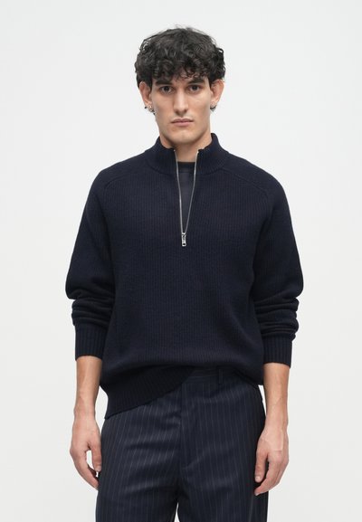 Navy ribbed knit pullover with a high collar and half zipper. The model pairs it with dark pinstriped trousers. Soft texture, relaxed fit.