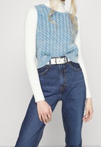 Top sin mangas de punto trenzado azul claro sobre un cuello de tortuga blanco con canalé, combinado con unos vaqueros de mezclilla oscuros de cintura alta y un cinturón blanco.