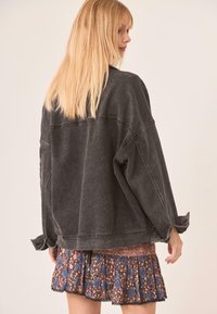 Mujer con cabello largo y rubio, vistiendo una chaqueta de mezclilla oscura y oversized y una falda corta floral con patrones en azul y rosa.