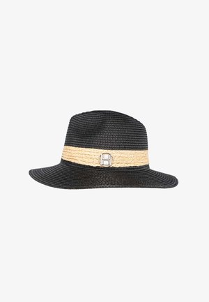 Kvinde smiler, iført beige strikket top med korte ærmer og shorts, justerer en sort og beige bredskyggede hat med en dekorativ nål.