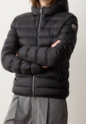 Henkilö, jolla on yllään musta tikattu toppatakki käsivarret ristissä, yhdistetty harmaisiin housuihin, vaaleaa taustaa vasten.