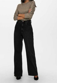 Schwarze weit geschnittene Jeans mit hoher Taille, ausgestattet mit fünf Taschen und subtiler Stickerei. Kombiniert mit einem hellbraunen strukturierten Pullover.