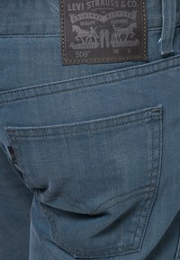 Siniset denimpellavahousut, joissa on teksturoitu pinta, takatasku ja musta nahkainen merkintä, jossa on kohopainetut brändäyksen yksityiskohdat.