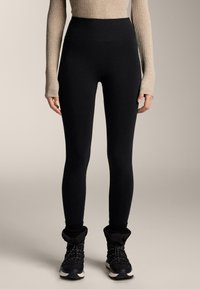Leggings noir taille haute, tissu extensible, texture lisse. Portés avec des bottines noires à chevilles avec des bordures tricotées et des lacets texturés.