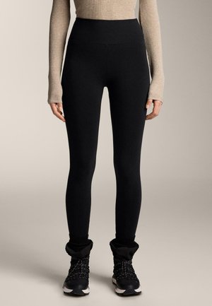 Leggings noir taille haute, tissu extensible, texture lisse. Portés avec des bottines noires à chevilles avec des bordures tricotées et des lacets texturés.