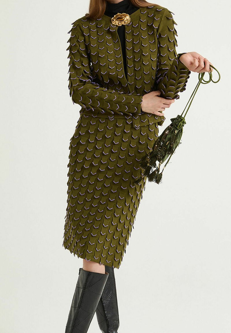 Robe vert olive à écailles texturées, accents violets et col montant. Associée à des bottes noires montantes et tenant un parapluie décoratif.