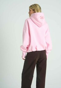 Růžová volná mikina s kapucí, pohodlným střihem a žebrovanými manžetami. Kombinováno s tmavě hnědými kalhotami s kapsami na zadní straně.