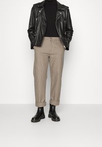 Veste de moto en cuir noir portée sur un col roulé noir, associée à un pantalon à carreaux beige texturé retroussé aux chevilles, et des bottes noires.