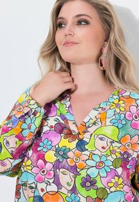La blusa colorata presenta un motivo floreale e facciale, con scollatura a V e maniche lunghe. Il materiale sembra leggero e morbido.
