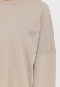 Sudadera beige con un acabado texturizado, que presenta un cuello redondo y un pequeño parche rectangular de logo en un tono más claro en el lado izquierdo del pecho.