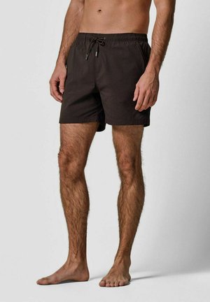 Man staand op blote voeten, gekleed in zwarte zwembroek met trekkoord, tegen een effen achtergrond.