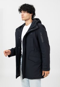 Schwarze gepolsterte Jacke mit Kapuze, ausgestattet mit einem vorderen Reißverschluss, mehreren Taschen und strukturierten Steppdetails. Über einem weißen Hemd getragen.