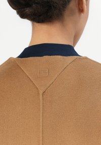 Bruine fleecejas met een gestructureerd oppervlak, voorzien van een marineblauwe geribbelde kraag en een subtiel logo dat op de achterkant is gedrukt.