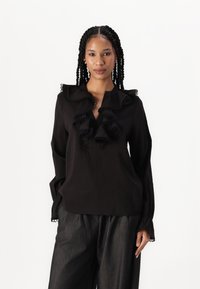 Blusa nera con scollatura a V, grande colletto volant e dettagli arricciati. Maniche lunghe con polsini gather. Abbinata a pantaloni scuri con texture.