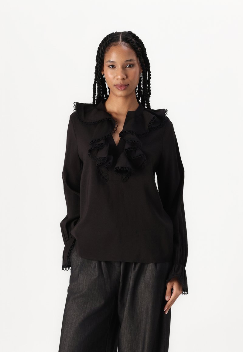 Blusa nera con scollatura a V, grande colletto volant e dettagli arricciati. Maniche lunghe con polsini gather. Abbinata a pantaloni scuri con texture.