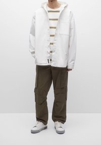 Chaqueta blanca de botones sobre una camisa a rayas en tonos beige y verde; pantalones cargo verde olivo con múltiples bolsillos; zapatillas blancas.