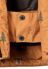 Orange tygjacka med inredning som har djur- och trädmönster. Svarta tryckknappar och kontrasterande fodertextur. Funktionella designelement.