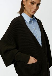 Tamno smeđi rebrasti kardigan s dubokim V-izrezom, prevelikim rukavima i bočnim džepovima, preko svijetloplavog košulje s kragnom.