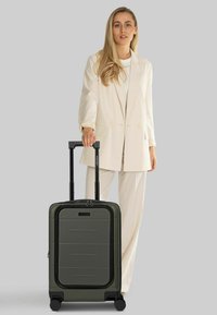 Donna in completo crema in piedi che tiene la maniglia di una valigia rigida verde oliva con quattro ruote su sfondo semplice.