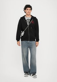 Hanorac negru cu fermoar, tricou gri cu grafică, blugi deschis la culoare, cu croială largă, geantă argintie, teniși alb-negru. Material neted, croială lejeră.
