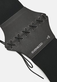 Black waist cincher with a faux leather front featuring lace-up detailing and grommets. The back is made of elasticated fabric for stretch.