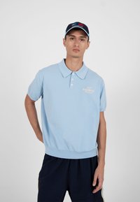 Jeune homme portant un polo bleu clair et un pantalon bleu marine avec une casquette assortie, debout devant un fond gris uni.