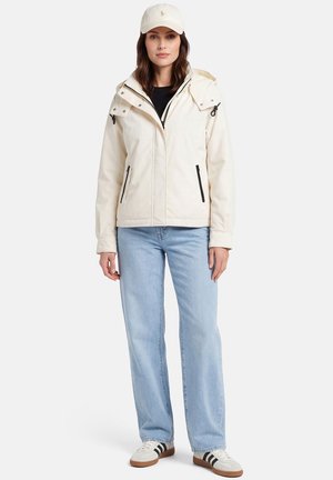 Vrouw die een off-white capuchonjas draagt, lichtblauwe spijkerbroek, witte pet en witte sneakers met zwarte strepen, staand tegen een effen achtergrond.