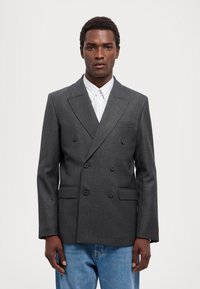 Blazer doppiopetto grigio scuro con rever a risvolto, due tasche frontali e una texture liscia, abbinato a una camicia a righe chiari.