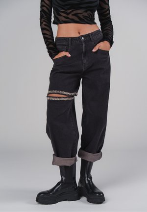 Person trägt schwarze, hochgeschnittene, weite Jeans mit Kristallketten an zerrissenen Oberschenkelstellen, schwarze klobige Stiefel und ein durchsichtig gestreiftes Langarmoberteil.