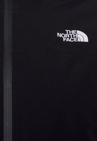 Svart jacka med dragkedjestängning, med en vit logotyp från The North Face på övre vänstra sidan. Slät tygtextur och minimalistisk design.