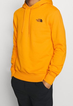 Personne portant un sweat à capuche de couleur orange vif The North Face avec cordons, les mains dans les poches d'un pantalon noir, sur un fond blanc uni.