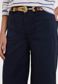 Tumsnēgbalti zili brīvā stilā bikšu, ar sānu kabatām un pinumu brūnas jostas ar sudraba sprādzi, novietotas uz gaiša fona.