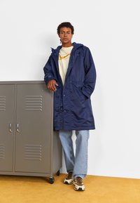 Parka impermeabile blu marina con cappuccio, dotata di tasche frontali, chiusura con zip e cuciture visibili, indossata sopra una maglietta leggera e jeans.