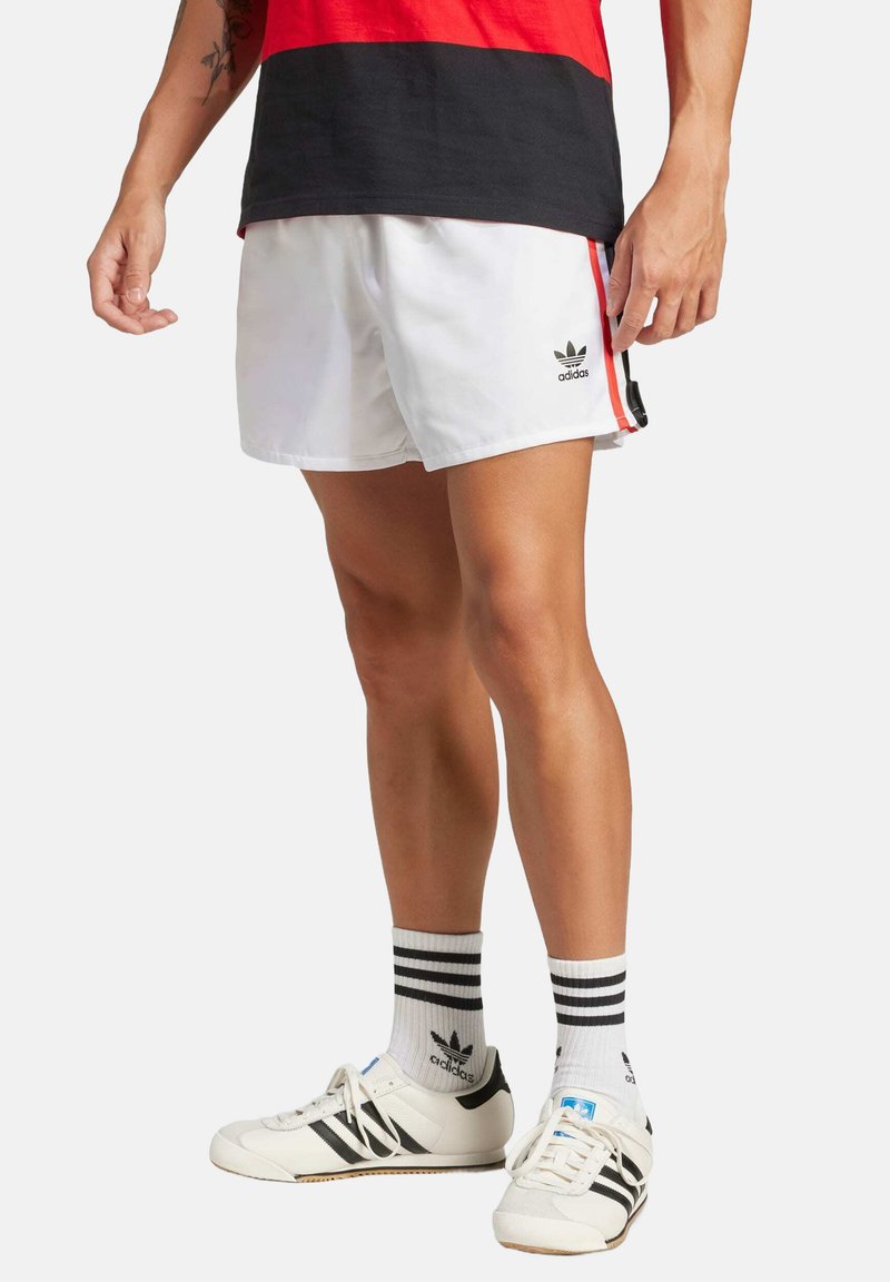 Bílé sportovní šortky s černým logem Adidas, doplněné černobílými pruhovanými ponožkami a černobílými botami, s elegantním designem.