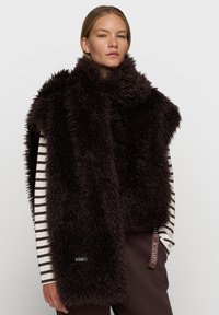 Sztuczny futrzany szal w ciemnym brązie o miękkiej, textured powierzchni; noszony na tle koszuli z długim rękawem w paski, z akcentami logo.