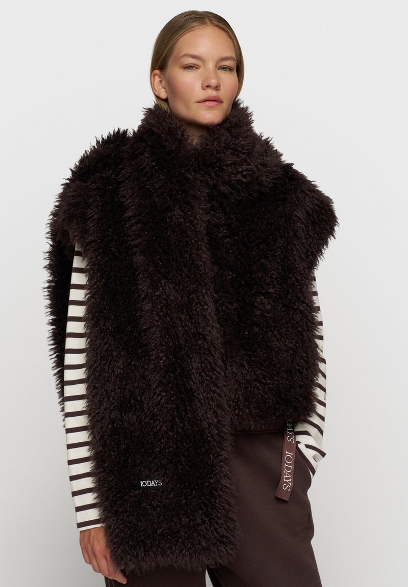 Sztuczny futrzany szal w ciemnym brązie o miękkiej, textured powierzchni; noszony na tle koszuli z długim rękawem w paski, z akcentami logo.
