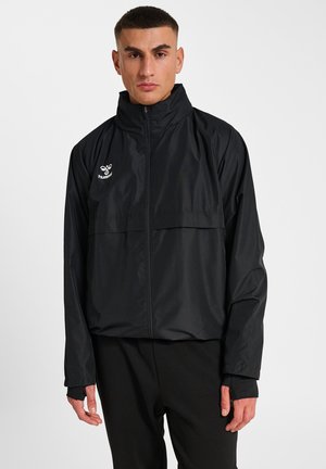 Un hombre posa con una chaqueta deportiva negra con cremallera y pantalones negros sobre un fondo liso y claro.