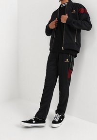 Henkilö, joka on pukeutunut mustaan Converse-harjoitussarjaan, jossa on punaisia ja vihreitä yksityiskohtia, sekä mustiin Converse-tennareihin, joissa on valkoiset pohjat ja nauhat.