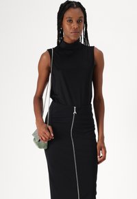TOP cu mânecă scurtă neagră și gât fals; fustă neagră ajustată cu fermoar pe lateral; modelul ține o geantă mică verde cu negru în carouri.