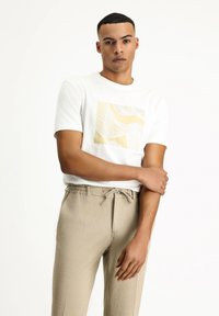 Camiseta de algodón blanca con un patrón abstracto amarillo en el frente, combinada con pantalones texturizados beige claro y una cinturilla elástica.