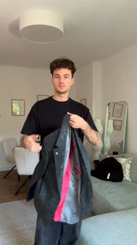 Sort jakke lavet af tekstureret stof med rød foring, der har en struktureret design og knapper. Baggrunden inkluderer møbler og indretning.