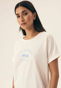 Biała bawełniana koszulka z niebieską grafiką z napisem "KOCHAJ SIEBIE" oraz dwiema ilustracjami chmur. Krótkie rękawy i luźny krój.
