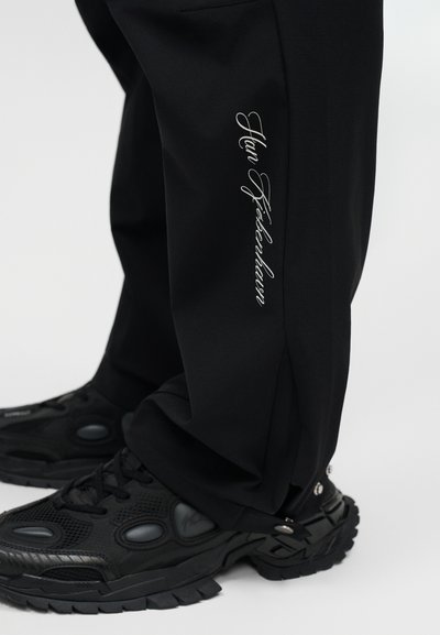 Κάτω μέρος πόδι που φορεί μαύρο παντελόνι με λευκό χειρόγραφο κείμενο "Han Kjøbenhavn" και μαύρα ογκώδη αθλητικά παπούτσια πάνω σε λευκό φόντο.