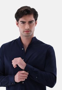 Marineblaues Langarmhemd mit Stehkragen, Knopfdetails an den Manschetten und einer glatten, glänzenden Textur. Sitzt eng am Körper.