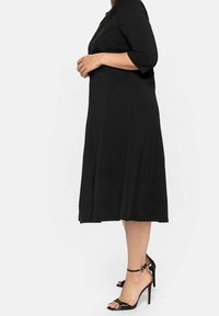Robe noire jusqu'au genou avec des manches trois-quarts, un corsage ajusté et une jupe évasée. Portée avec des sandales à talons hauts noires.